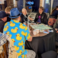 A group of people sat around a table talking. One person wearing a brightly coloured shirt with yellow ducks on.